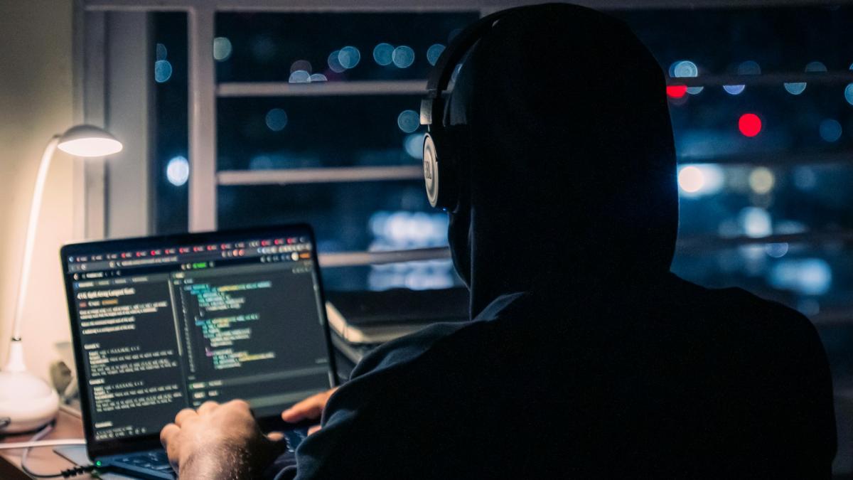 Хакер в капюшоне сидит перед монитором с кодом